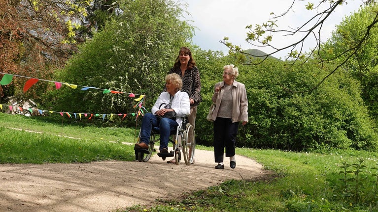 Three people on the accessible path at Ilam Park. One person is in a wheelchair and the others are walking alongside her. They are smiling and chatting.